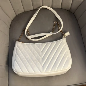 Vit quiltad axelväska med guldkedja - Snygg vit axelväska från Glitter med quiltad design och guldfärgad kedjedetalj på axelremmen. Väskan har dragkedja upptill och ett rymligt fack med innerficka. Perfekt för dig som vill ha en stilren och trendig accessoar. Den har lite rost på kedjan samt lite missfärgad.