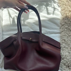 Vinröd handväska i skinn med gulddetalj - Stilren vinröd handväska i skinn med två rejäla handtag och snygg gulddetalj framtill. Väskan har rymligt svart foder och innerfack med dragkedja. Perfekt för dig som vill ha en klassisk och elegant look med plats för allt du behöver. Notera att väskan är ifrån Shein, köpte den för 167 kronor. 