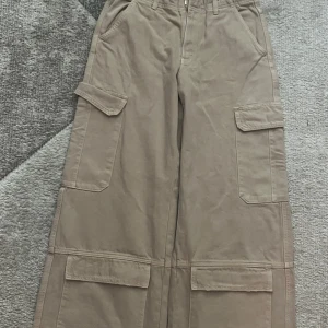 Beiga cargopants från Zara, strl 36 - Säljer ett par beiga cargopants från Zara i storlek 36. Byxorna har bred passform, stora fickor på sidorna och raka ben. Tillverkade i bomull och perfekta för en chill och trendig look.