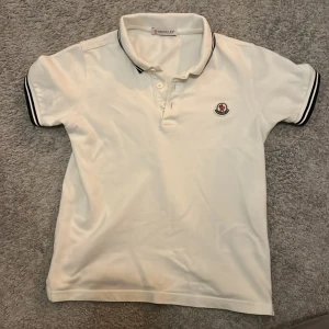 Vit Moncler pikétröja med logga - Vit pikétröja från Moncler med klassisk krage och knäppning framtill. Svarta ränder på krage och ärmslut ger en sportig vibe. Broderad Moncler-logga på bröstet. Tillverkad i bomull för en skön och luftig känsla.