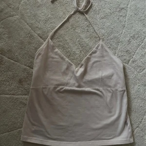 Ljusbeige halterneck linne - Supersöt ljusbeige linne med halterneck och knyt i nacken. Toppen har v-ringad skärning och ärmlös design, perfekt för varma dagar. Mjuk bomullskänsla och enkel, stilren look som passar till jeans eller kjol.