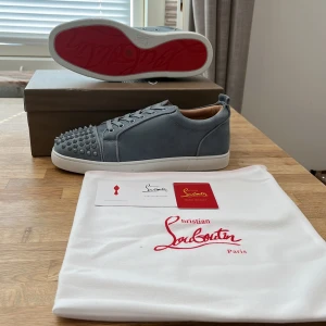 Blå Christian Louboutin sneakers med nitar - Snygga blå sneakers från Christian Louboutin i mocka med ikoniska röda sulor. Skorna har rund tå, snörning och coola nitar framtill som ger en edgy vibe. Insidan är beige och sulan är vit med röd undersida. Perfekta för dig som vill sticka ut.
