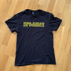 Svart AIK t-shirt Norra Ståplats - Svart t-shirt med gul text 'Norra Ståplats AIK Stockholm' på bröstet och stort Aik-emblem på ryggen. Klassisk rund hals och korta ärmar. Materialet känns mjukt och skönt mot huden. 