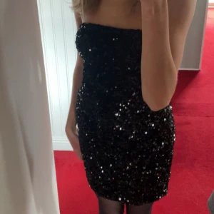 Svart glittrig tubklänning - Säljer en svart, glittrig kortklänning med paljetter. Klänningen är axelbandslös och har en tight passform som framhäver figuren. Perfekt för dig som vill sticka ut och glänsa på festen!