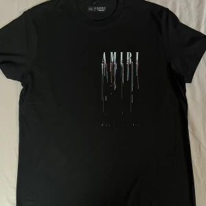 Svart AMIRI t-shirt med ängeltryck - Svart t-shirt från AMIRI med coolt tryck framtill där loggan droppar i olika färger och stor vit ängelgrafik på ryggen. Klassisk passform och rund halsringning. Perfekt statement-plagg för dig som gillar streetwear vibes.