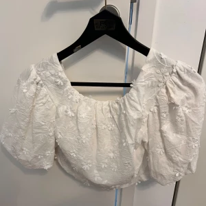 Vit broderad blus från NA-KD - Supersöt vit blus från NA-KD med puffiga korta ärmar och är lite kortare i längden. Toppens tyg är broderat med blommor och har en luftig känsla, perfekt för varma dagar. Passar dig som gillar romantisk och trendig stil.