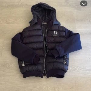 Svart pufferjacka från UrbanEdge - Snygg svart pufferjacka från UrbanEdge med huva och silvriga dragkedjor. Jackan har ribbade muddar, quiltad kropp och släta ärmar. Perfekt för kalla dagar och har en clean streetstyle-vibe. Logga på bröstet för extra detalj.