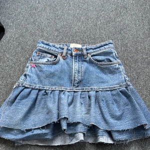 Volangkjol i jeans från Neuw - Supersöt blå jeanskjol från Neuw med volang och råa kanter. Klassisk femficksmodell, liten rosa rosett vid fickan och snyggt läderpatch bak. Storlek 24, passar en 170 som highwaist men passar också 146/152 som lowwaist. Kan anpassa priset.