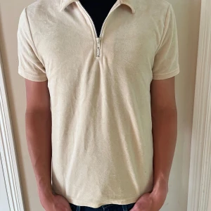 Beige pikétröja med half zip - Snygg beige pikétröja med kort ärm och krage. Tröjan har en stilren half zip-dragkedja framtill och är gjord i ett mjukt, frottéliknande material. Perfekt för dig som gillar en clean och avslappnad look.