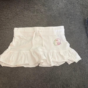 Vit volangkjol Paradise Beach - Supersöt vit kortkjol med volangkant och snörning i midjan. Tryck med färgglad text och palm på sidan där det står Paradise Beach. Perfekt för sommaren. Pris kan diskuteras 