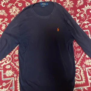 Mörkblå tröja Polo Ralph Lauren S - Mörkblå långärmad tröja från Polo Ralph Lauren i slim fit. Tillverkad i mjuk pima bomull och har den klassiska orange loggan broderad på bröstet. Enkel och stilren design med rund halsringning och ribbade muddar.