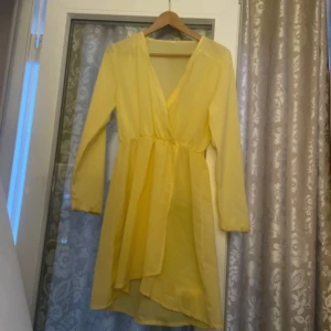 Gul omlottklänning med lång ärm - Snygg gul omlottklänning med lång ärm och v-ringning. Klänningen har en lätt och luftig känsla, markerad midja och asymmetrisk nederdel. Perfekt för dig som vill sticka ut med en färgglad look.