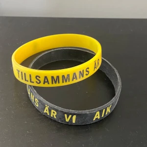 AIK armband i gult och svart - Två silikonarmband i AIK:s färger, ett gult med svart text och ett svart med gul text. Texten lyder 'Tillsammans är vi AIK'. Perfekt accessoar för dig som vill visa ditt AIK-stöd.