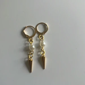 Pearly spike  - Snygga örhängen i guldton med rund vit pärla och en cool konformad detalj längst ner. Hängande modell med små ringar som ger en trendig vibe. Perfekta för dig som vill sticka ut med en mix av klassiskt och edgy.