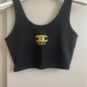 Svart croppad linnetopp - Svart croppad linnetopp från med guldig broderad logga och text framtill. Toppen har breda axelband och rundad halsringning, perfekt att styla med höga jeans eller kjol. Mjuk och stretchig bomullskänsla.