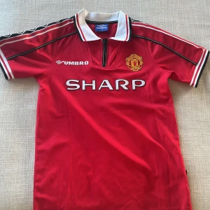 Manchester United Beckham 7 Umbro tröja - Klassisk Manchester United fotbollströja från Umbro med Beckham 7 på ryggen. Röd med vita och svarta detaljer, broderat klubbmärke och SHARP-tryck på bröstet. Kortärmad modell i lätt och ventilerande material, perfekt för dig som älskar retrostil och fotbollshistoria.