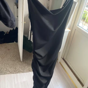 Svart långklänning med drapering - Säljer en svart långklänning med smala axelband och snygg drapering längs sidorna. Klänningen har en elegant cowlneck-ringning och är figurnära med en stilren look. Perfekt för dig som vill ha en klassisk och tidlös stil.