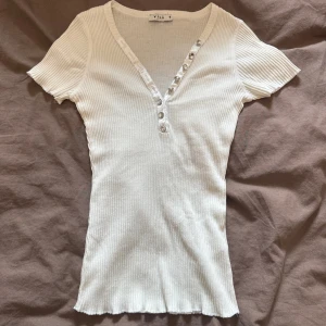 Vit ribbad topp med knappar från Vila - Supersnygg vit ribbad topp från Vila med korta ärmar och v-ringning. ❤️😍Dekorativa silvriga knappar framtill och glansig kant längs knappraden ger en extra touch. Figurnära passform och stretchigt material som sitter skönt. Några stenar är borta men inget som riktigt syns(se mer på bild 2)! ❤️