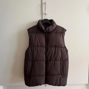Brun dunväst från Monki - Säljer en brun, quiltad dunväst från Monki i storlek S. Västen har hög krage, dragkedja framtill och två fickor på sidorna. Perfekt för lager-på-lager och chill höst- eller vinteroutfits.