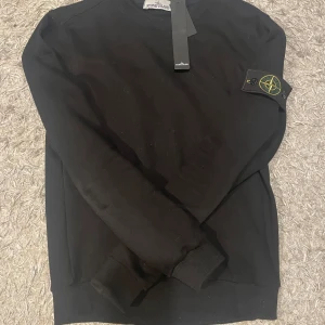Svart sweatshirt från Stone Island - Svart sweatshirt från Stone Island med klassisk rund hals och ribbade muddar. Ikonisk logotyp-patch på vänster ärm. Tröjan är i mjuk bomull och har en clean, stilren look som passar perfekt till streetwear-stilen. Pris kan diskuteras vid snabb affär.