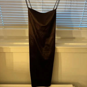 Brun satinklänning med tunna axelband - Snygg brun kort satinklänning med tunna dubbla axelband och öppen rygg med knyt. Klänningen har en figurnära passform och en lätt glansig yta som ger en lyxig känsla. Perfekt för dig som vill sticka ut med en elegant look. vissa ytliga märken och skiftningar i tyget finns