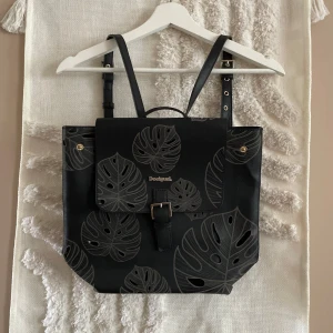 Svart ryggsäck med blad från Desigual - Snygg svart ryggsäck från Desigual med grått bladmotiv och justerbara axelremmar. Väskan har ett spänne framtill, dragkedja på baksidan med guldfärgad S-detalj och är gjord i syntetmaterial. Perfekt för dig som vill sticka ut med en unik och trendig accessoar.