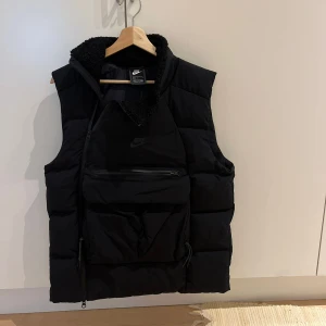 Nike tech väst Svart  - Säljer en svart dunväst från Nike med stor ficka framtill och dragkedja. Västen har hög krage med mjukt foder och diskret Nike-logga på bröstet. Perfekt för lager-på-lager och streetwear-stil.