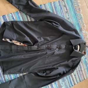 Svart skjorta från Burberry - Snygg svart skjorta från Burberry med klassisk krage och knappar framtill. Insidan av knappslån har det ikoniska rutiga Burberry-mönstret i beige, svart och rött. Skjortan är långärmad och i ett slätt bomullstyg, perfekt för en clean och stilren look.