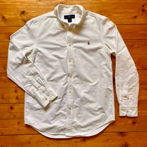 Vit skjorta från Ralph Lauren - Klassisk vit skjorta från Ralph Lauren med button down-krage och broderad logga på bröstet. Skjortan har långa ärmar, knappar framtill och är tillverkad i mjuk bomull. Perfekt för en clean och stilren look. Storleken säger 18 men jag skulle säga att den passar en Small