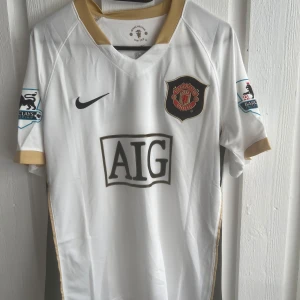 Manchester United Ronaldo 7 bortatröja - Vit Manchester United bortatröja med guldiga detaljer, AIG sponsortryck och klubbmärke på bröstet. Premier League-patch på ärmarna och Ronaldo 7-tryck på ryggen. Tillverkad i lätt och ventilerande material, perfekt för fotbollsfans.