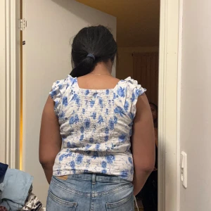 Blommig volangtopp från Lindex - Supersöt topp från Lindex i vitt med blåa blommor, volangdetaljer på axlarna och smockad resår över hela toppen. Ärmlös modell med rundad halsringning, perfekt för en fräsch och somrig look. Materialet är mjukt och stretchigt för extra komfort.             STÅR EJ FÖR POSTENS SLARV💘.                   INGA RETURER TACK💗🌸💞💕