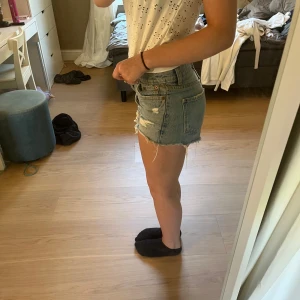 Blå jeansshorts med slitningar - Säljer ett par klassiska blå jeansshorts med hög midja och råa, fransiga kanter. Shortsen har slitna detaljer framtill och bakfickor. Perfekta för sommaren och ger en avslappnad vibe till din outfit.