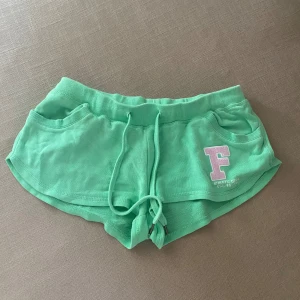 Gröna mjuka shorts med snörning - Snygga gröna shorts med bred resår i midjan och snörning framtill. Mjukt material och två fickor. På vänster sida finns en vit och rosa F-applikation med texten 'ATHLETIC DEPT'. Perfekta för chill eller träning.