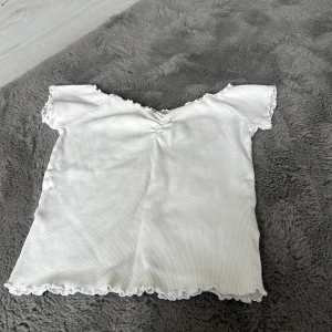 Vit ribbad topp från Mix - Supersöt vit ribbad topp från Mix med korta ärmar och v-ringning. Toppen har volangkant vid hals, ärmslut och nederkant samt en lätt rynkning framtill. Perfekt för dig som gillar en feminin och trendig stil.
