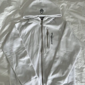 Vit hoodiejacka från Sail Racing - Vit hoodiejacka från Sail Racing med dragkedja framtill och en extra ficka med dragkedja på bröstet. Jackan har broderad logga på huvan och bröstet. Tillverkad i mjukt bomullsmaterial och har långa ärmar. Perfekt för en clean och sportig look.