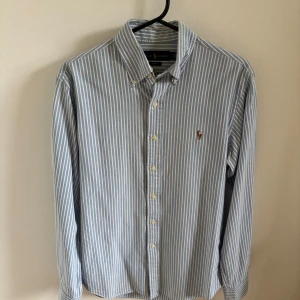 Blåvit randig skjorta Ralph Lauren - Snygg blå och vit randig skjorta från Ralph Lauren i slim fit-modell. Skjortan har klassisk krage, knäppning framtill och broderad logga på bröstet. Tillverkad i mjuk bomull och har långa ärmar med manschettknappar. Använd ett fåtal gånger.