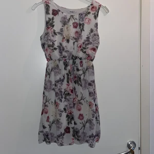 Blommig sommarklänning i pastell - Söt ärmlös kortklänning från New Look med blommigt mönster i lila, rosa och grå toner. Klänningen har markerad midja och luftig passform, perfekt för varma dagar. Tunn och lätt i materialet, vilket ger en skön känsla.