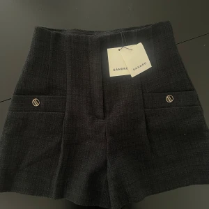 Svarta högmidjade shorts från Sandro - Stilrena svarta högmidjade shorts från Sandro i strukturerat tyg med två fickor framtill och snygga guldfärgade knappar med logga. Klassisk rak passform och exklusiv känsla, perfekta för en trendig look.