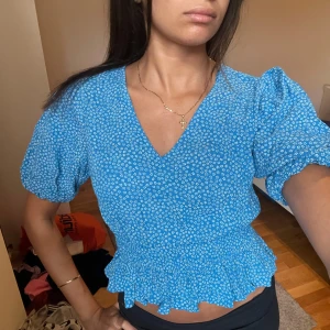 Blommig blå blus från BikBok - Supersöt blå blus från BikBok med små vita blommor, v-ringning och puffiga korta ärmar. Smockad midja med volangkant ger en feminin och trendig look. Perfekt till jeans eller kjol för en fräsch stil.