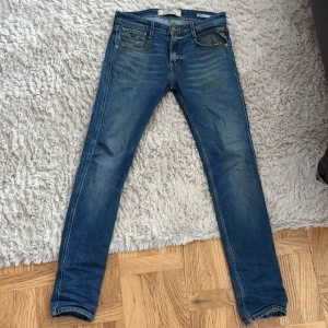 Replay Anbass blå jeans slim fit - Snygga blå Replay Anbass jeans med slim fit och klassisk femficksdesign. Jeansen har ljusa slitningar framtill, läderpatch med Replay-logga bak och kontrastsömmar. Perfekta för dig som gillar en modern och stilren look. 7/10 cond, Små slitningar som detaljer.