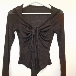 Svart glittrig långärmad body med snörning - Svart långärmad topp i glittrigt material med djup v-ringning och snygg snörning framtill som ger en rynkad effekt.
