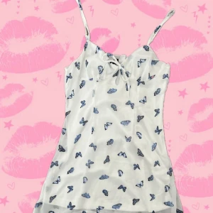 Vit kortklänning med blå fjärilar H&M - Supersöt vit kortklänning från H&M Divided med blå fjärilsmönster, smala axelband och liten rosett framtill. Klänningen har dragkedja i sidan och är tillverkad i ett lätt, luftigt material som passar perfekt för varma dagar.