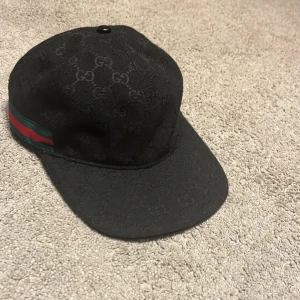 Svart Keps - Snygg svart keps med klassiskt GG-monogram över hela tyget. Kepsen har en grön och röd rand på sidan och justerbar rem baktill. Tillverkad i bomull och polyester, perfekt för dig som vill sticka ut med en lyxig accessoar., Knappast Använd använd para för test osv