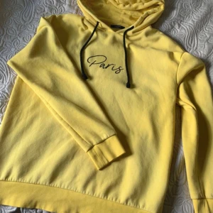 Gul hoodie från Jack & Jones Paris - Gul hoodie från Jack & Jones med svart snörning och texten 'Paris' tryckt på bröstet. Mjuk insida och klassisk huva gör den perfekt för chill dagar. Oversized känsla och långa ärmar. Grym färg som sticker ut och passar dig som gillar streetstyle.