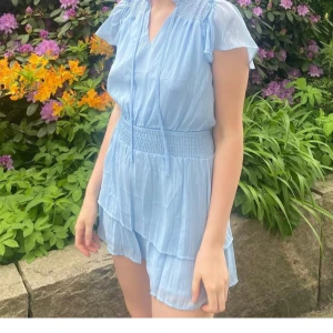 Ljusblå kortklänning med volang - Supersöt ljusblå kortklänning med v-ringning, smock i midjan och volangdetaljer. Klänningen har korta ärmar och tunna knytband vid halsen. Perfekt för varma dagar och ger en fräsch sommarkänsla.