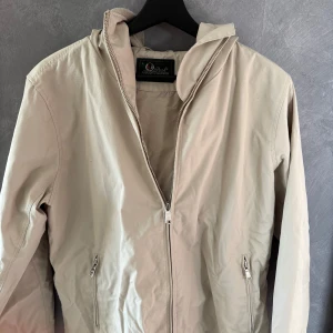 Beige vindjacka med huva från Peak Performance - Snygg beige vindjacka från Peak Performance med huva och dragkedja framtill. Jackan har två fickor med dragkedja och är tillverkad i ett lätt bomullsmaterial. Perfekt för blåsiga dagar och enkel att matcha med olika outfits.