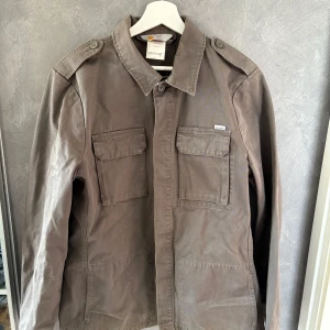 Brun overshirt från Carhartt, L - Säljer en brun overshirt från Carhartt i 100% bomull. Jackan har två stora bröstfickor med lock, axelklaffar med knapp och klassisk krage. Perfekt för lager-på-lager och enkel att styla till jeans eller cargobyxor.