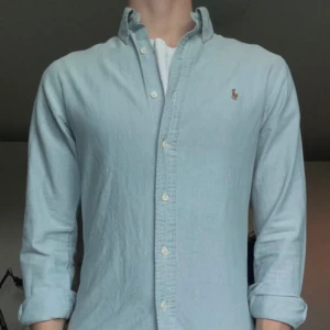 Ljusblå skjorta från Ralph Lauren - Klassisk ljusblå skjorta från Ralph Lauren med button-down krage och broderad logga på bröstet. Skjortan har långa ärmar, knappar framtill och är tillverkad i mjuk bomull. Perfekt för en clean och stilren look.