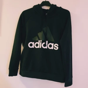 Svart Adidas hoodie med logga strl S - Svart hoodie från Adidas med stor vit logga framtill och huva med snörning. Tröjan har långa ärmar och är tillverkad i mjuk bomullsblandning. Perfekt för en sportig och avslappnad stil.  Helt skick dock ev lite ljusare i färgen & en obehandlad fett fläck