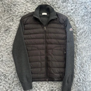 Grå Moncler pufferjacka i ull och dun - Snygg grå pufferjacka från Moncler med quiltad framsida i dun och stickade ärmar och rygg i ull. Jackan har dragkedja framtill, hög krage och Moncler-logga på ärmen. Perfekt för dig som vill ha en stilren och exklusiv look.
